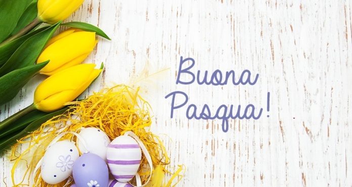Uova di Pasqua di colore viola e bianco disposte in un nido di rafia gialla decorativa. A fianco alcuni tulipani gialli su uno sfondo chiaro. Al centro della foto la scritta Buona Pasq